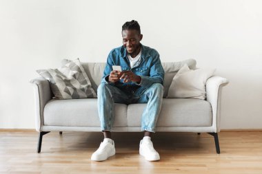 Happy Black Male Using Phone Texting And Browsing Internet Sitting On Couch At Home. Millennial Guy Using New Mobile App Via Cellphone. Gadgets And Technology Concept