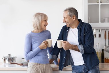 Beautiful loving senior couple in homewear standing at white cozy kitchen, drinking fresh morning coffee together, enjoying good morning, sharing plans for day, laughing, copy space