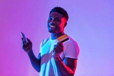 Portrait of cheerful young black man holding cellphone and credit card, using banking or shopping mobile app, receiving cashback, trading or gambling online in neon light