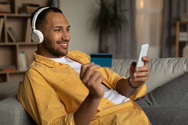 Remote payment, online order concept. Happy young black man in headphones buying things on web, using smartphone and credit card, enjoying shopping in internet at home