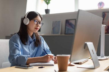 Asian Businesswoman Working On Computer And Taking Notes Sitting At Desk In Modern Office Indoors. Female Manager Works Online At Workplace Wearing Eyeglasses And Headphones. Side View