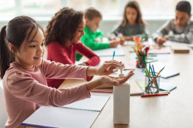 Covid 19 Protection. Asian Schoolgirl Disinfecting Hands With Sanitizer Sitting Learning With Multiethnic Classmates In Modern Classroom Indoor. Healthcare And School Education In Pandemic