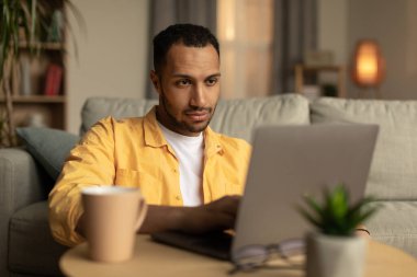 Serious millennial black male using laptop pc at home office. Handsome young man using portable computer, working or studying online, having remote job, watching video indoors
