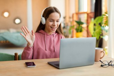 Positive teenage girl cheerfully waving at laptop screen, using wireless headphones and smiling. Smiling school girl having video chat with tutor, enjoying studying from home
