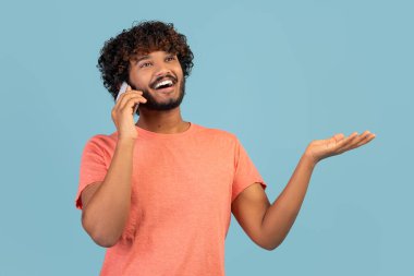 Closeup of happy millennial hindu guy with curly hair having phone conversation with someone on blue studio background, gesturing, sharing exciting news, looking at copy space and smiling