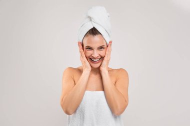 Bath Fun. Cheerful Middle Aged Woman Wrapped In Towel Posing Over Light Grey Background, Happy Joyful Mature Female Touchinbg Face And Laughing At Camera, Enjoying Beauty Care Routine, Copy Space