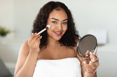 Daily makeup. Beautiful oversize black lady applying blush with makeup brush, holding mirror and looking at camera. Young woman using new cosmetics while getting ready at home