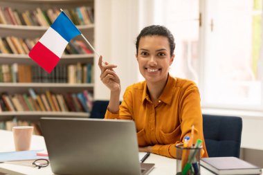 Cheerful mixed race female teacher giving French class, communicating on laptop, sitting in library interior. Experienced college professor giving new material to students on web