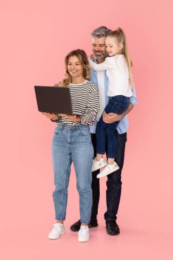 Cheerful Parents And Kid Daughter Using Laptop Browsing Internet Together Over Pink Studio Background. Family Advertising Website Holding Computer. Vertical Shot