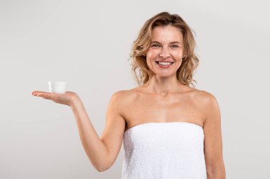 Anti-Age Cosmetics. Beautiful Middle Aged Female Holding Jar With Face Cream On Her Palm, Smiling Attractive Lady Wrapped In Towel After Bath Posing On Light Grey Studio Background, Copy Space