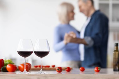 Selective focus on two glasses of red wine on kitchen table over dancing senior couple, romantic elderly husband and wife cooking healthy dinner together, celebrating marriage anniversary