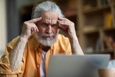 Stressed senior man freelancer working from home, sitting on couch, using laptop, touching his head, having troubles at job, suffering from migraine, headache, copy space, closeup shot