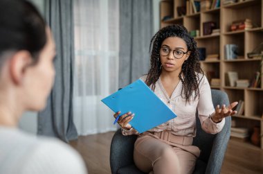 Serious african american young woman psychologist advises european lady patient in clinic interior. Mental problems, professional medical care, fight against depression, work with client due covid-19 clipart