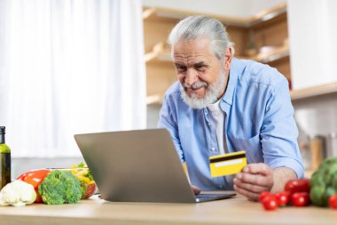 Smiling Senior Man Using Laptop And Credit Card While Cooking Food In Kitchen, Happy Elderly Male Ordering Groceries Online, Making Internet Purchases With Computer At Home, Copy Space