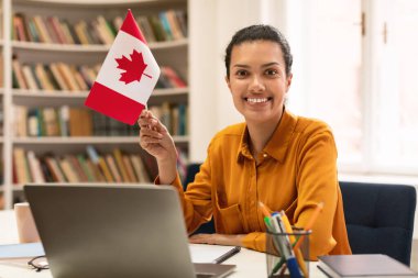 Happy mixed race woman holding flag of Canada, using laptop computer and smiling at camera, sitting in library. Modern online foreign education, emigration and citizenship, working abroad concept