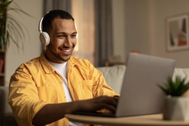 Joyful young African American man using laptop, having remote job, wearing headphones at home. Confident millennial black male working on pc, typing on keyboard, surfing web indoors