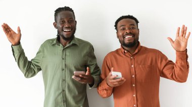 Hello. Two Cheerful Black Guy Using Smartphones And Waving Hands Smiling To Camera Posing Together Standing Over White Studio Background. Mobile Offer And Application Advert Concept. Panorama
