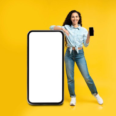 Great App. Arabic Female Leaning On Big Cellphone With Blank Screen Showing Smartphone And Advertising Mobile Application Standing Over Yellow Studio Background. Square, Mockup