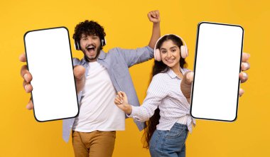 Cheerful Indian Couple Wearing Headphones Holding Big Blank Smartphones With White Screen, Happy Young Man And Woman Recommending New Online Music Streaming Service, Mockup, Creative Collage