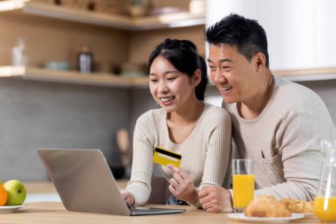 Cute japanese family shopping from home while having lunch together, happy chinese middle aged man and young woman sitting at kitchen table, drinking juice, using laptop and credit card, empty space
