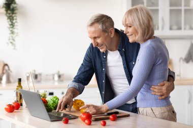 Loving senior couple watching online food vlog on Internet while cooking together at home, positive senior man and woman cutting vegetables and looking at computer screen, copy space, kitchen interior