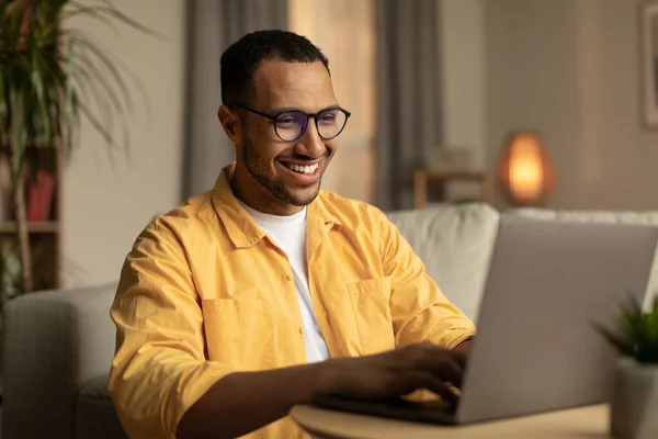 Beautiful young black man working on laptop pc, enjoying remote job or education in living room. Smiling millennial guy using portable pc for work or online studies, communicating on web indoors