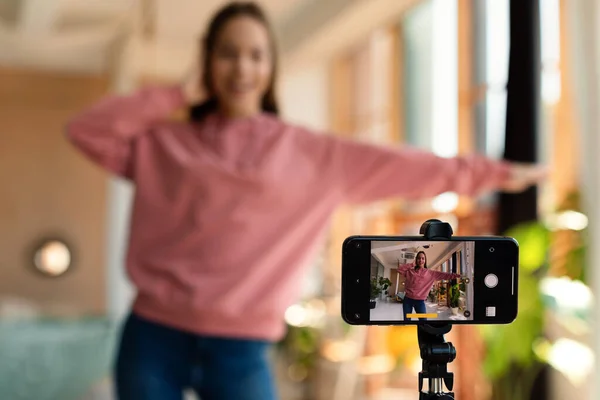 Modern teenage female influencer recording video on smartphone, dancing on camera at home, selective focus. Teen lifestyle vlogger making content for her blog, filming for social media