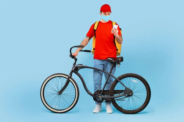 Food Delivery App. Courier Guy Wearing Face Mask Using Cellphone Standing With Bike Delivering Meal Over Blue Studio Background. Transportation Service And Offer Concept