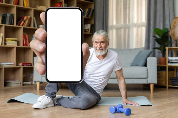 Cheerful handsome athletic senior man sitting on yoga mat, showing modern cell phone with white empty screen, recommending nice fitness mobile application, home interior, copy space, mockup