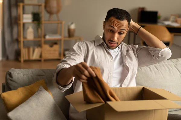 Dissatisfied buyer concept. Frustrated black man unboxing package after delivery, holding clothes, sitting on couch at home, copy space. African American guy unhappy about online purchase