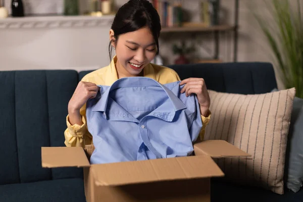 Shopping And Delivery. Joyful Chinese Lady Unpacking Cardboard Box Holding Blue Shirt Receiving Parcel From Shop Sitting On Couch At Home. Ecommerce Service, Contented Customer Concept