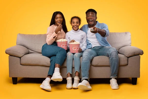 Overjoyed african american family watching movie and eating popcorn from buckets, parents sitting with daughter on couch, man holding remote control, yellow background