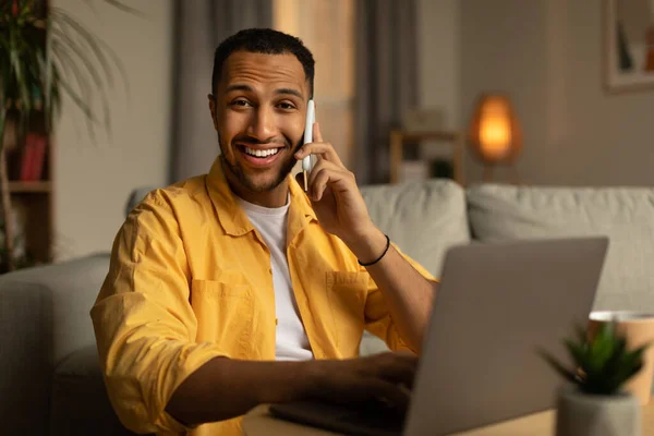 Handsome young black man in casual wear talking on smartphone, enjoying conversation at home office. Attractive African American guy speaking with customer while working on laptop in living room