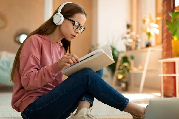 Distance education concept. Smart teen girl sitting on floor with book, doing home assignment, wearing headphones, listening online lesson and reading at home