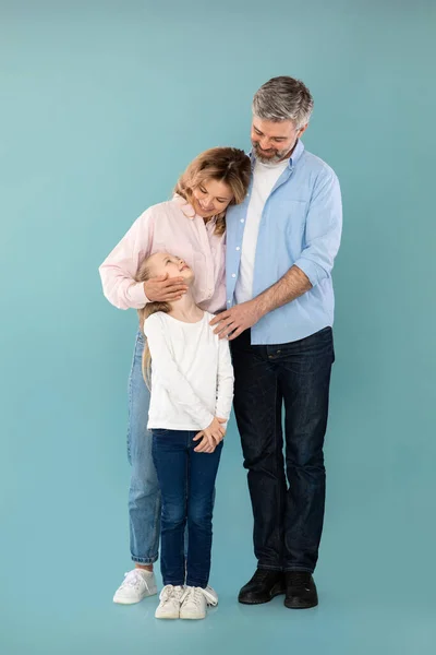 Vertical Shot Of Cheerful Family Embracing Expressing Love Posing Together Over Blue Background In Studio. Mature Parents Hugging Standing With Kid Daughter. Full Length
