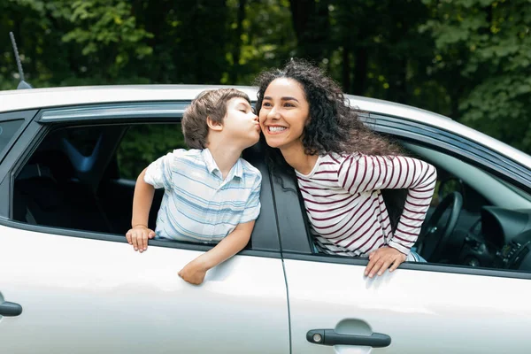 Cheerful little arabic boy kisses his mother on cheek through open window in new car, enjoy journey together, outdoor. Summer trip with family, love and family relationship, auto tour at vacation