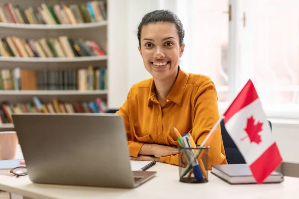 Online foreign languages tutoring. Happy female teacher sitting in library with flag of Canada, communicating on laptop with students and giving new material on web