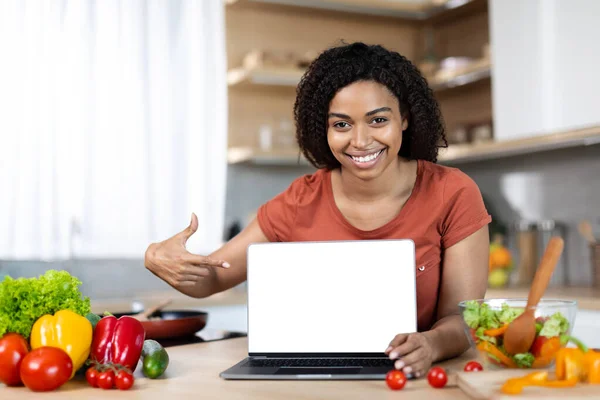 Smiling young african american woman blogger housewife pointing finger at laptop with blank screen in cozy kitchen interior with organic vegetables. Food blog, chat, great website, ad and huge offer