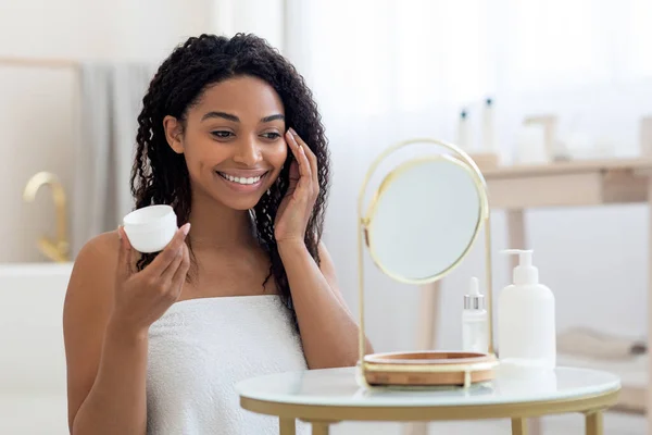 Attractive Young African American Woman Applying Moisturising Cream On Face, Beautiful Black Lady Wrapped In Towel Looking At Mirror And Holding Jar With Anti-Aging Cosmetic Product, Copy Space
