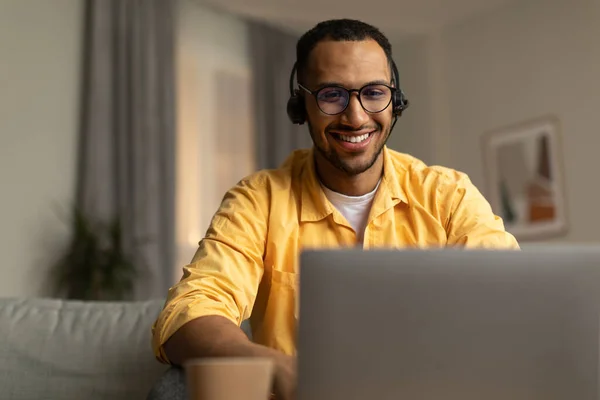 Smiling young black man in wireless headphones working on laptop, watching webninar in living room, copy space. Cheerful African American male freelancing, taking part in online conference