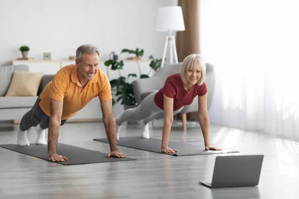 Athletic senior spouses training at home, smiling mature man and woman planking together, using modern laptop, watching fitness class on Internet, exercising at home, panorama with copy space