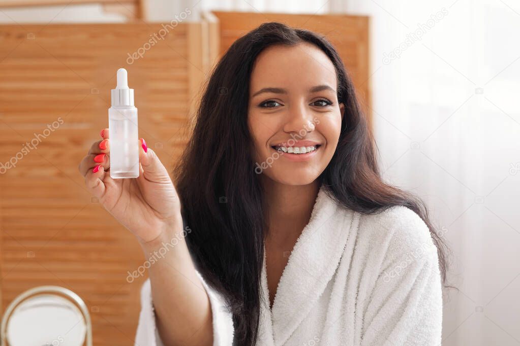 Pretty Woman Showing Cosmetic Bottle Recommending Serum Posing In Modern Bathroom At Home. Female Advertising Facial Skin Care Products Smiling To Camera Indoors. Skincare Concept