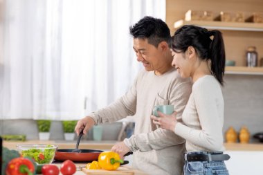 Loving middle aged chinese man husband cooking delicious healthy dinner for his beautiful young wife, happy asian spouses having conversation while preparing food at kitchen, copy space