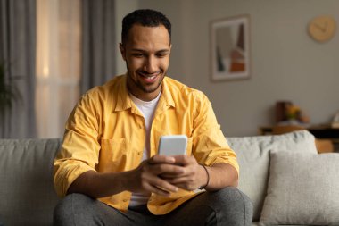 Joyful young black guy using smartphone, video chatting with friend, browsing social media, sitting on sofa at home, copy space. Millennial African American man with cellphone surfing web indoors
