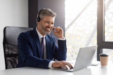 Middle aged businessman with headset and laptop making video conference with international partners while sitting at workplace in office, smiling male entrepreneur enjoying online communication