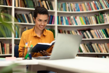 Virtual lecture. Happy mixed race woman sitting in library, holding book, having online class with tutor and looking at laptop computer. Woman studying remotely