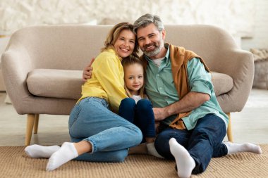 Happy Parents And Kid Daughter Hugging Sitting On Floor Smiling To Camera Posing At Home. Loving Family Of Three Embracing Spending Time Together. Happiness Concept