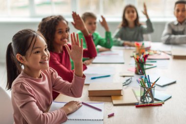 Happy Multiethnic School Kids Learning Raising Arms During Lesson Sitting At Desk In Classroom At School. Modern Education And Knowledge Concept. Selective Focus