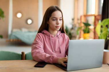 Portrait of female teenager sitting at desk and using laptop computer at home, typing on keyboard. Focused youth looking at screen, doing homework or making video call with teacher