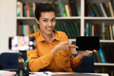 E-Learning concept. Cheerful lady tutor having video conference using cellphone on tripod, showing digital tablet with blank screen, recording tutorial with mobile phone, streaming live to students